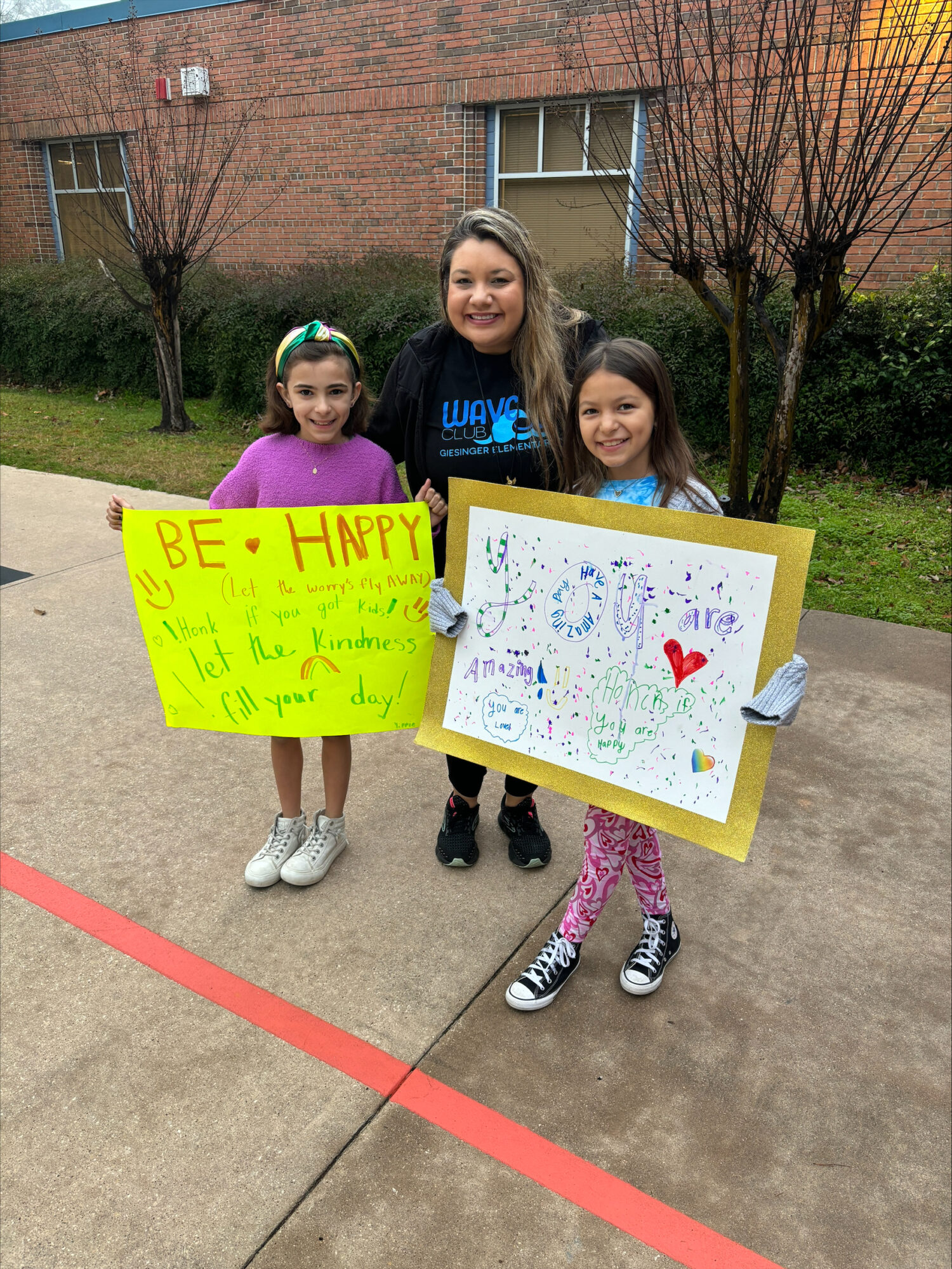 Two girls and their teacher hold up signs to greet people in the car line as members of the Geisinger Wave Club.